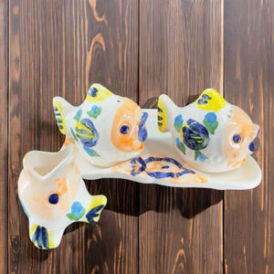 May include: A set of three ceramic salt and pepper shakers shaped like fish. The shakers are painted with colorful floral designs and have a white background. The shakers are sitting on a white ceramic tray.