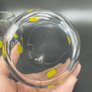 May include: Clear glass tumbler with yellow polka dots. The glass has a rounded shape and is partially filled. The yellow dots are evenly spaced around the glass. The glass is held by a person.