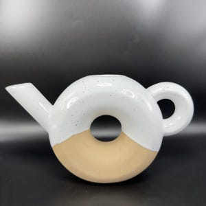 Peut inclure: Une th&eacute;i&egrave;re en c&eacute;ramique de forme originale en forme de donut. La moiti&eacute; sup&eacute;rieure est blanche avec des d&eacute;tails mouchet&eacute;s, tandis que la moiti&eacute; inf&eacute;rieure est beige. Elle poss&egrave;de un bec verseur et une anse arrondie.