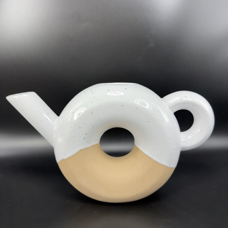 Peut inclure: Une th&eacute;i&egrave;re en c&eacute;ramique de forme originale en forme de donut. La moiti&eacute; sup&eacute;rieure est blanche avec des d&eacute;tails mouchet&eacute;s, tandis que la moiti&eacute; inf&eacute;rieure est beige. Elle poss&egrave;de un bec verseur et une anse arrondie.