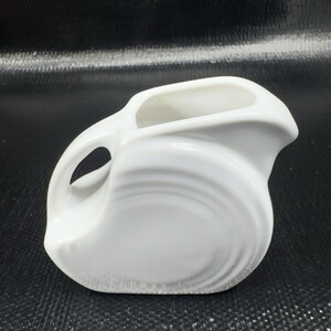 May include: A white ceramic pitcher with a unique, curved design. The pitcher features a handle with a circular opening and a spout. The body of the pitcher has a swirling pattern. The pitcher is likely used for serving beverages.