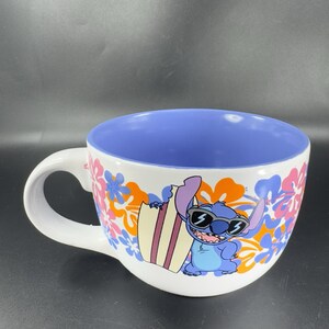 May include: White ceramic mug with a blue interior. The mug features a cartoon image of Stitch from Lilo & Stitch wearing sunglasses and holding a surfboard. The background is a colorful floral pattern.