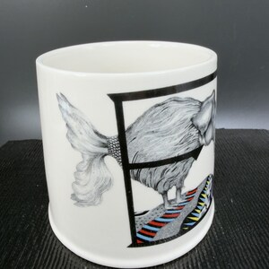 May include: White ceramic mug featuring a black letter "F" framing a detailed illustration of a fish-like creature. The creature has a textured, feathery body and a patterned tail. The base of the design includes colorful stripes.