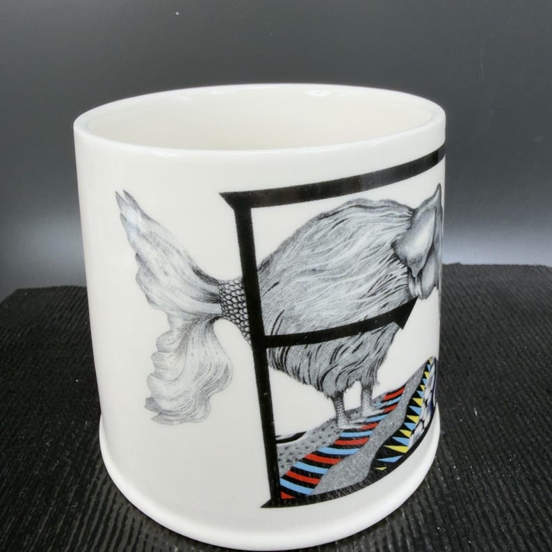 May include: White ceramic mug featuring a black letter "F" framing a detailed illustration of a fish-like creature. The creature has a textured, feathery body and a patterned tail. The base of the design includes colorful stripes.