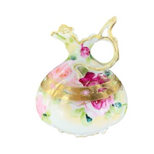 May include: A white porcelain pitcher with a floral design. The pitcher has a wide, round body and a short, curved spout. The floral design features pink and red roses with green leaves. The pitcher is decorated with a gold band around the middle.