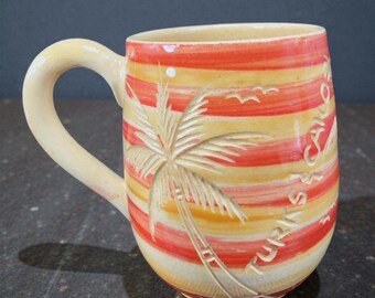 May include: Ceramic mug with a cream-colored handle and a design featuring a palm tree and the words "Turks & Caicos." The mug has a striped pattern in shades of yellow, orange, and red, with a carved palm tree design.