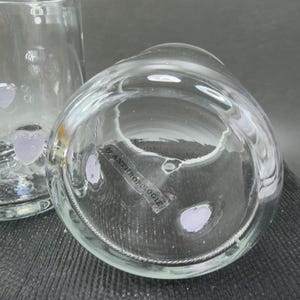 May include: Clear glass vase with a rounded shape, featuring a label that reads "by ANTHROPOLOGIE". The vase has a textured surface and a decorative design. The image shows the bottom of the vase.