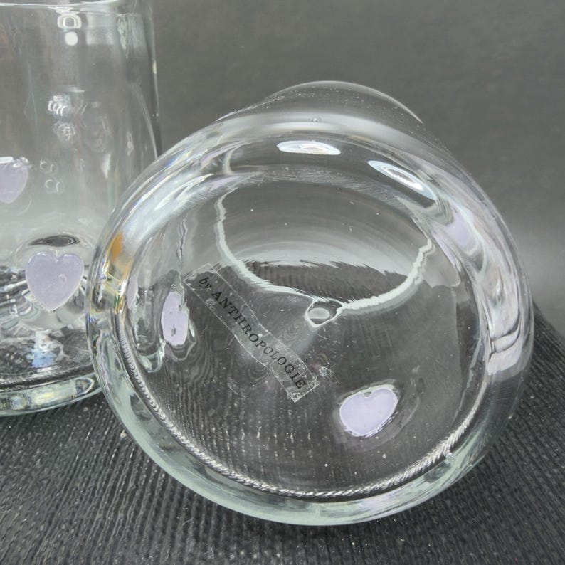 May include: Clear glass vase with a rounded shape, featuring a label that reads "by ANTHROPOLOGIE". The vase has a textured surface and a decorative design. The image shows the bottom of the vase.