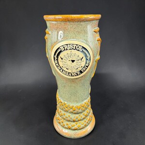 May include: A tall, ceramic mug with a green and orange glaze. The mug features a circular emblem with the text "Bristol Renaissance Faire" and a sun design. The base has a textured, scale-like pattern.