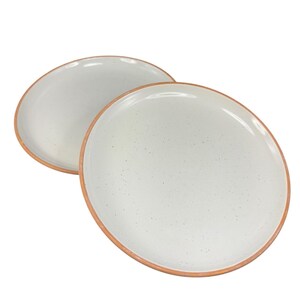 May include: Two off-white ceramic plates with speckled surfaces and a warm brown rim. The plates are round and slightly angled, suggesting a modern, minimalist design. Ideal for serving meals.
