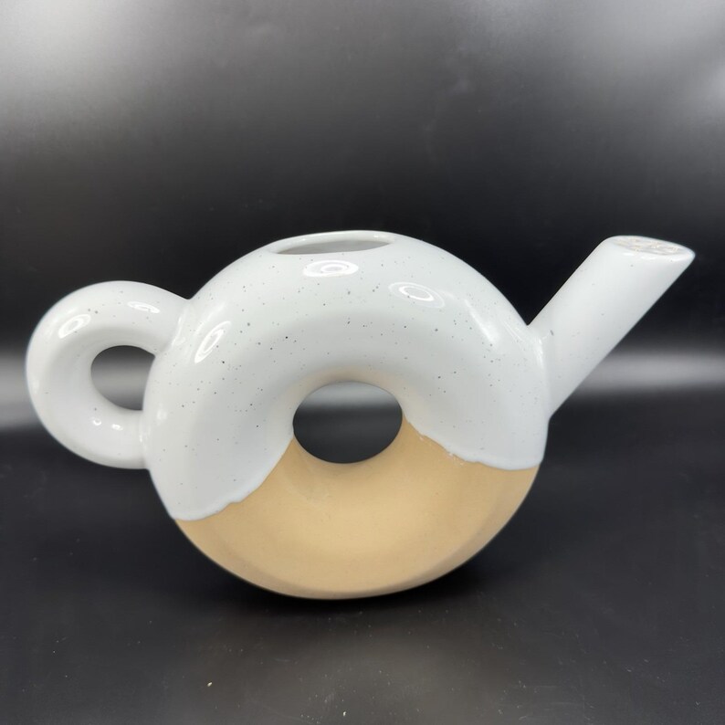 Peut inclure: Une th&eacute;i&egrave;re en c&eacute;ramique de forme originale en donut. La moiti&eacute; sup&eacute;rieure est blanche avec des d&eacute;tails mouchet&eacute;s, la moiti&eacute; inf&eacute;rieure est beige clair. Elle poss&egrave;de une anse arrondie et un bec verseur, id&eacute;ale pour le th&eacute;.