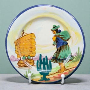 May include: Hand-painted ceramic plate with a blue rim, featuring a scene of a person carrying a bag, a yellow structure, and a cactus. The plate has a white background with yellow and pink accents, and is held by two white hooks.