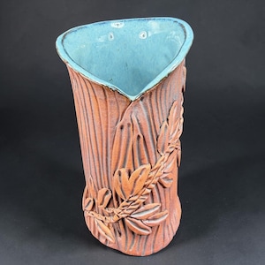 May include: A ceramic vase with a textured, reddish-brown exterior featuring a raised leaf and vine relief pattern. The interior of the vase is glazed in a smooth, bright turquoise blue, contrasting with the earthy, matte-finished outer surface.