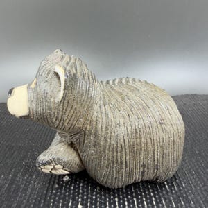 May include: A gray bear figurine with a textured surface. The bear is in a seated position, with its paws and head visible. The bear's snout is a light beige color, and the eyes and nose are black.