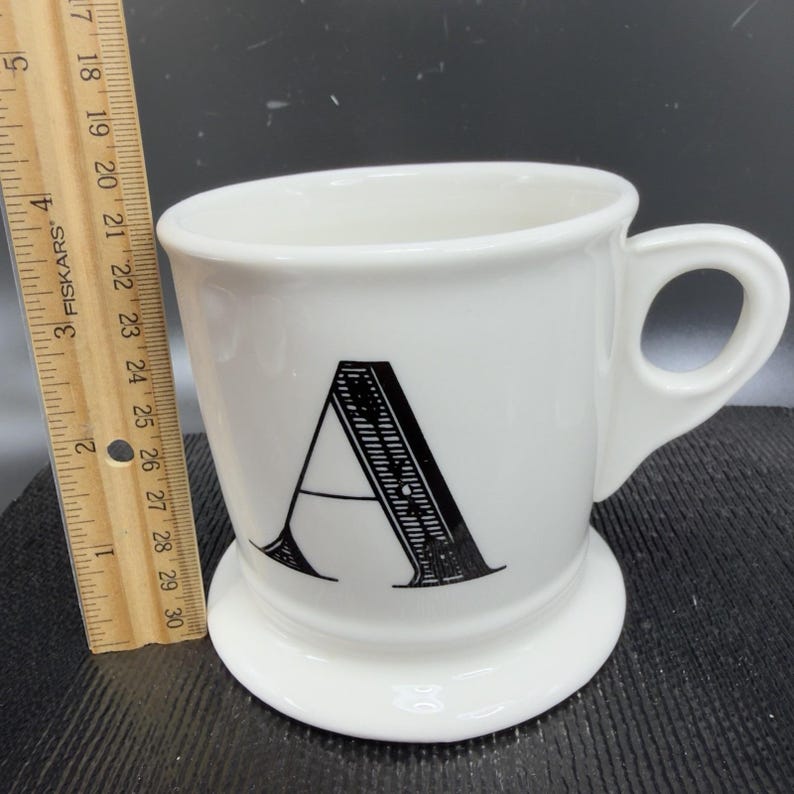May include: White ceramic mug with a black letter "A" on the front. The mug has a handle and a wide base. A ruler is next to the mug, showing measurements in inches.