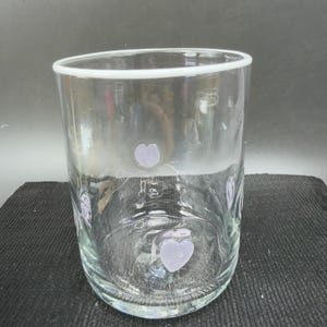 May include: A clear glass tumbler with a white rim and lavender heart-shaped designs. The glass is cylindrical and appears to be empty. The hearts are evenly spaced around the glass.