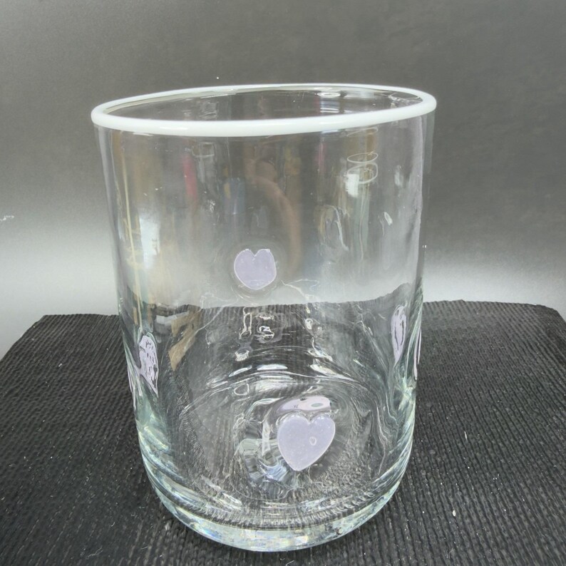 May include: A clear glass tumbler with a white rim and lavender heart-shaped designs. The glass is cylindrical and appears to be empty. The hearts are evenly spaced around the glass.
