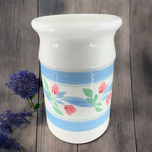 May include: White ceramic utensil holder with a blue and green floral design. The design features pink flowers and green leaves.