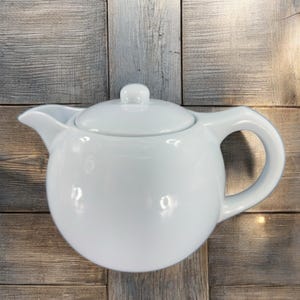 May include: A white ceramic teapot with a round body and a lid. The teapot has a spout and a handle.