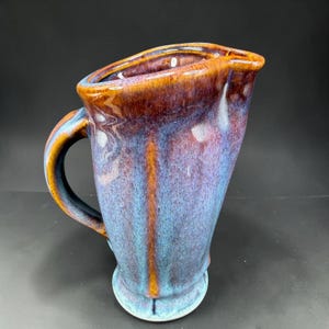 May include: A ceramic pitcher featuring a glossy glaze with blended hues of deep brown, violet, and light blue. The vessel has a curved handle, a wide pouring spout, and a vertical ribbed texture that runs along the body of the pitcher.