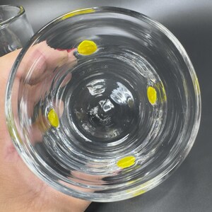 May include: Clear glass drinking glass with three yellow dots inside. The glass is round with a slightly flared rim. The glass is held by a hand, and the background is dark.