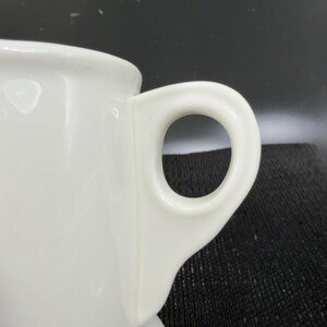 May include: Close-up of a white ceramic mug with a rounded handle. The mug is glossy and appears to be clean. The handle is a simple, circular design, and the mug's body is smooth. The background is dark.