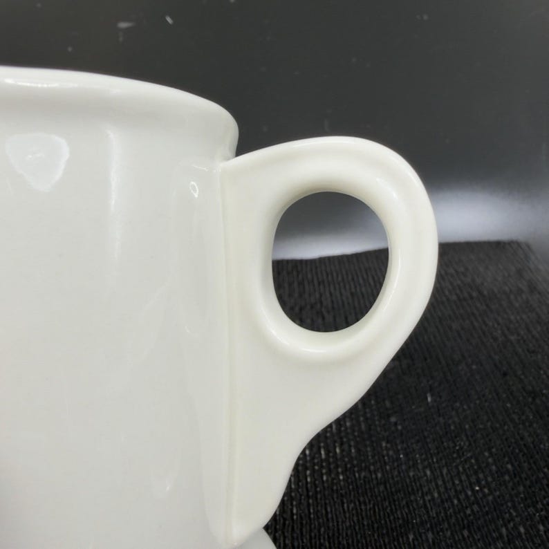 May include: Close-up of a white ceramic mug with a rounded handle. The mug is glossy and appears to be clean. The handle is a simple, circular design, and the mug's body is smooth. The background is dark.