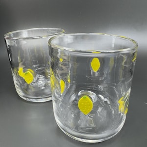 May include: Two clear, thick-walled drinking glasses with yellow lemon designs. The glasses are cylindrical with a slightly textured surface. The lemons are painted in a bright yellow color and are scattered around the glass.