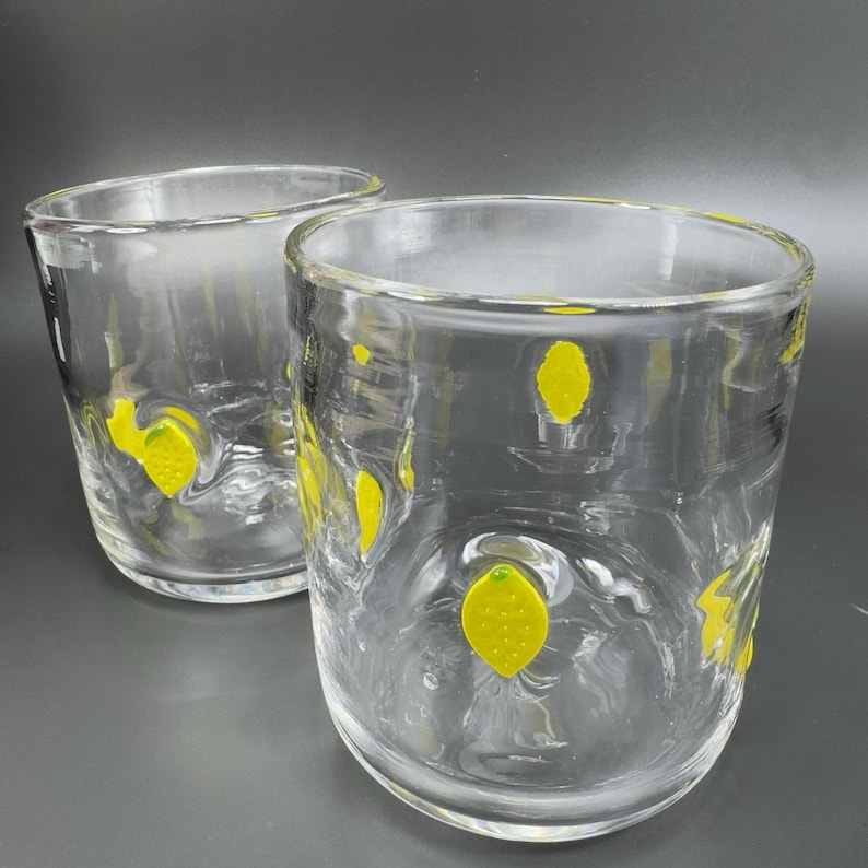 May include: Two clear, thick-walled drinking glasses with yellow lemon designs. The glasses are cylindrical with a slightly textured surface. The lemons are painted in a bright yellow color and are scattered around the glass.