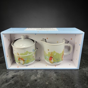 May include: A Peter Rabbit sugar and creamer set in a light blue box. The ceramic pieces feature illustrations of Peter Rabbit in a meadow scene. The sugar bowl has a lid, and the creamer has a handle and spout. The box is decorated with polka dots.