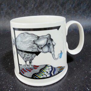 May include: White ceramic mug featuring a whimsical illustration of a gray elephant with a black outline. The elephant is standing on a colorful, patterned base. A small blue figure waters the elephant. The mug has a curved handle.
