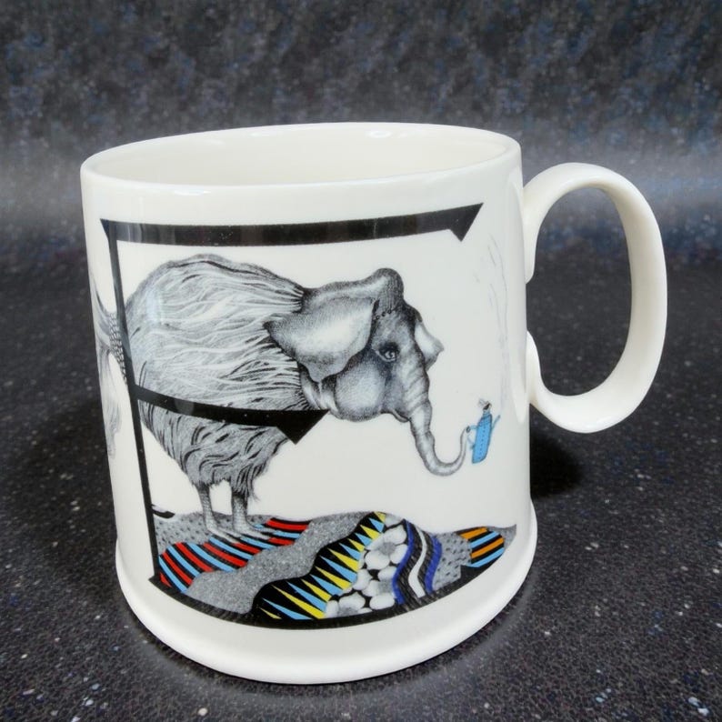 May include: White ceramic mug featuring a whimsical illustration of a gray elephant with a black outline. The elephant is standing on a colorful, patterned base. A small blue figure waters the elephant. The mug has a curved handle.