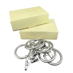 May include: Two cream-colored boxes with the text "Pottery Barn New York" and "set of seven" are stacked. Below are seven silver metal curtain rings with attached clips, designed for hanging curtains. The rings are circular and have a matte finish.