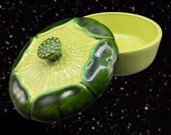 May include: A green and yellow ceramic dish with a lid. The lid has a flower design with a green knob in the center. The dish is round and has a smooth, glossy finish.