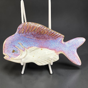 May include: A fish-shaped ceramic dish featuring a speckled glaze in shades of purple, blue, and white. The upper portion displays a textured, iridescent purple and blue finish, while the lower body is a smooth, off-white with subtle speckles.