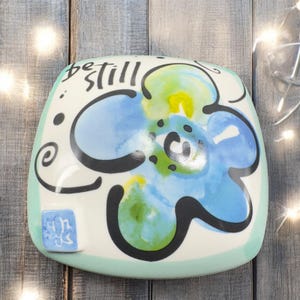 May include: Square ceramic dish with a hand-painted blue flower design and the words "be still" in black script. The dish has a light blue border and a small square emblem. Decorative item.