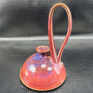 May include: A handmade ceramic spoon rest with a vibrant red and blue glaze. The spoon rest features a rounded base, a small circular holder, and a tall, arched handle. The glaze has a speckled appearance, adding visual interest to the piece.