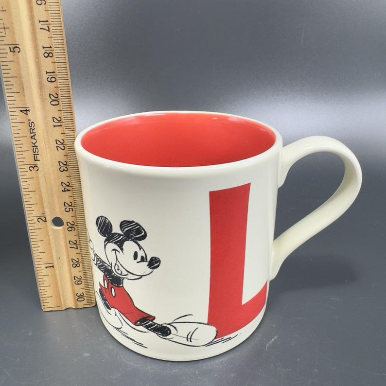 May include: A white ceramic mug with a red interior and a large red letter "L". The mug features a black and red illustration of Mickey Mouse. The mug has a curved handle.