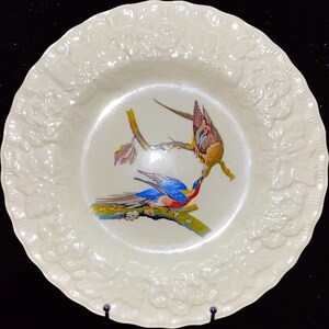 May include: A white ceramic plate with a raised floral design around the edge. The center of the plate features a painted image of two birds perched on a branch.
