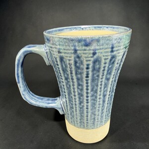 May include: A handmade ceramic mug with a blue and white textured design. The mug has a light beige base and a curved handle. The interior of the mug is a light yellow color. The mug is approximately 4 inches tall.