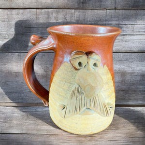 May include: A brown and tan ceramic mug with a face sculpted on the front. The face has a large nose, two eyes, and a mustache.
