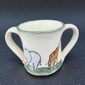 May include: A white ceramic mug with two handles, featuring hand-painted illustrations of an elephant and a giraffe on a green grassy base. The rim is trimmed with a green border. A charming, whimsical design.