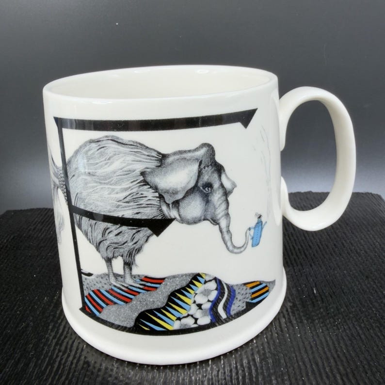 May include: White ceramic mug featuring a detailed illustration of a gray elephant with a blue watering can. The elephant stands on a colorful, patterned base, framed by a black border. The mug has a curved handle.