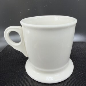 May include: A white ceramic mug with a rounded handle and a wide base. The mug has a simple, classic design, perfect for coffee or tea. The mug is made of a glossy, smooth ceramic material.