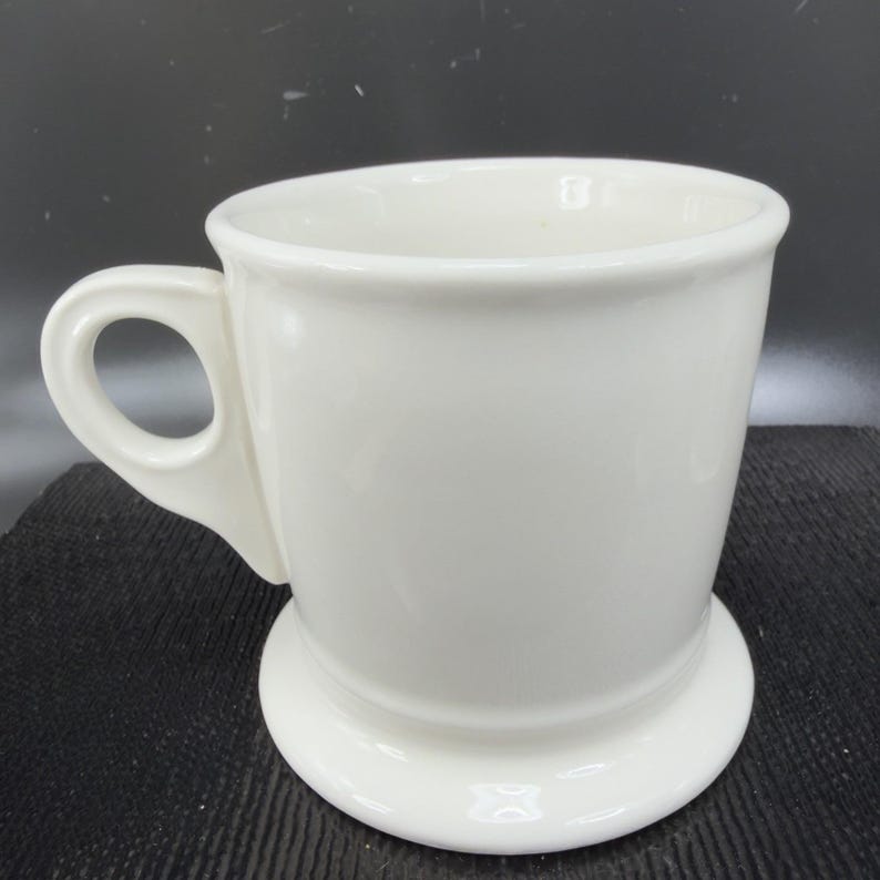 May include: A white ceramic mug with a rounded handle and a wide base. The mug has a simple, classic design, perfect for coffee or tea. The mug is made of a glossy, smooth ceramic material.
