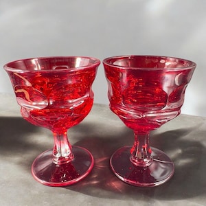 May include: Two red glass wine goblets with a textured design. The goblets have a round bowl and a tall stem with a round base.