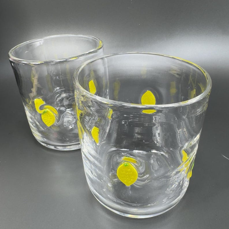 May include: Two clear glass tumblers with yellow lemon designs. The glasses are cylindrical with a slightly curved shape. The lemons are painted on the outside of the glasses. The background is a dark gray.