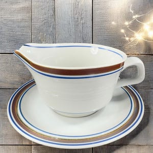 May include: White ceramic gravy boat with a matching saucer. The gravy boat has a brown and blue stripe design around the rim and a handle. The saucer has a matching brown and blue stripe design. A classic addition to any dining table.