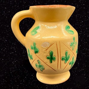 May include: A yellow ceramic pitcher with a green and brown clover design. The pitcher has a wide mouth and a handle.