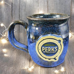 May include: A handcrafted blue ceramic mug with a large handle. The mug features a glossy finish and a cream-colored emblem with the words "PERKS of Frankfort" and the words "Coffee, Tea, Espresso, Fresh Baked Goods".